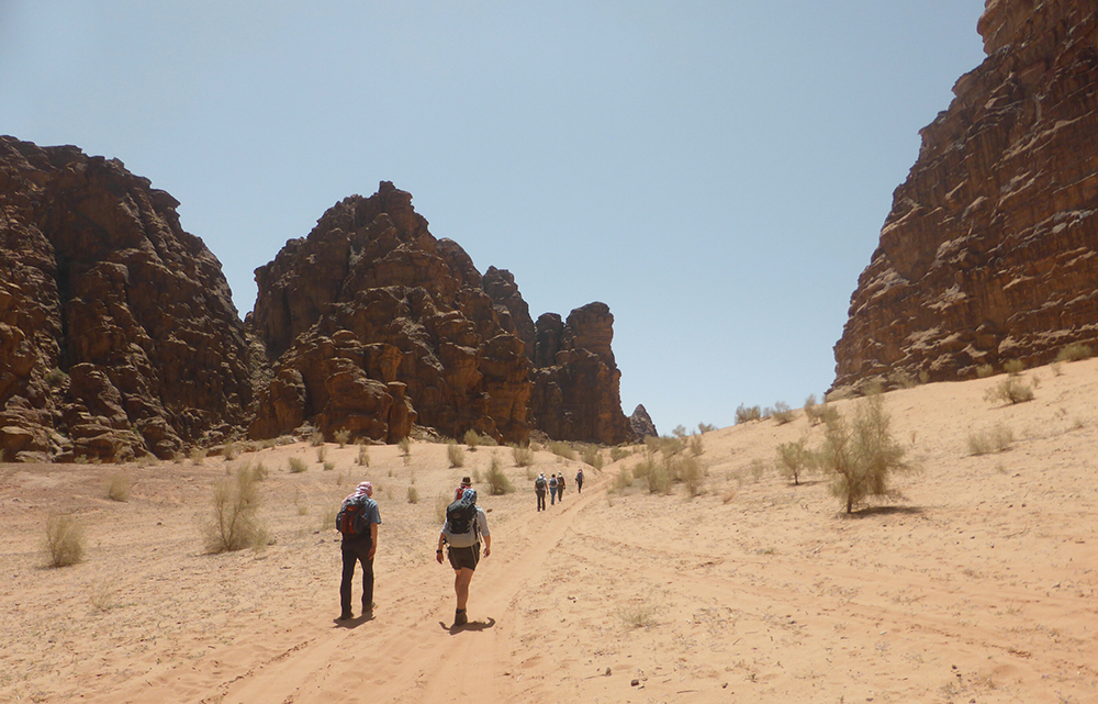 Hikers walk along a sandy path between tall, rugged cliffs in a sunlit desert landscape, with sparse vegetation scattered around.
